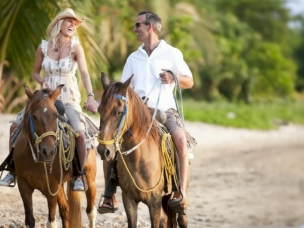 A couple is happily horseback riding on a sandy beach, surrounded by lush greenery and palm trees under a clear sky.