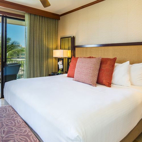 A cozy hotel room features a large bed with pillows, a bench at the foot, lamps, and a balcony view with palm trees outside.