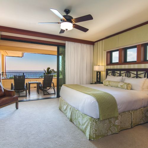 A hotel room with a king-sized bed, ceiling fan, and balcony view. Outdoor seating overlooks a scenic ocean view.
