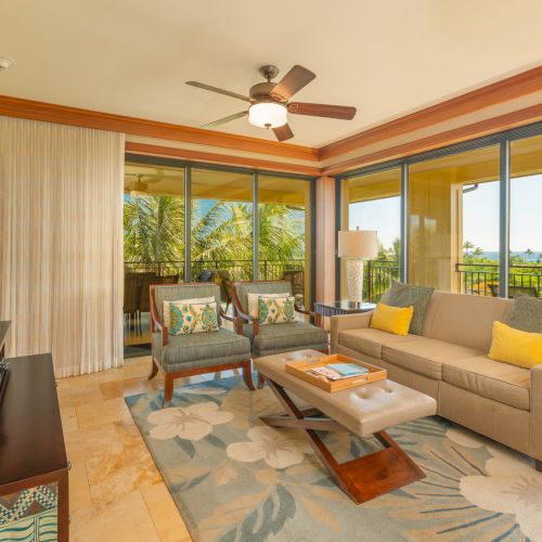 A cozy living room features a sofa, chairs, a table, and a TV. Large windows open to a balcony with a view. Light decor completes the space.