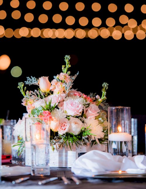 A beautifully set table with a floral centerpiece, surrounded by candles and glassware, under soft glowing string lights.