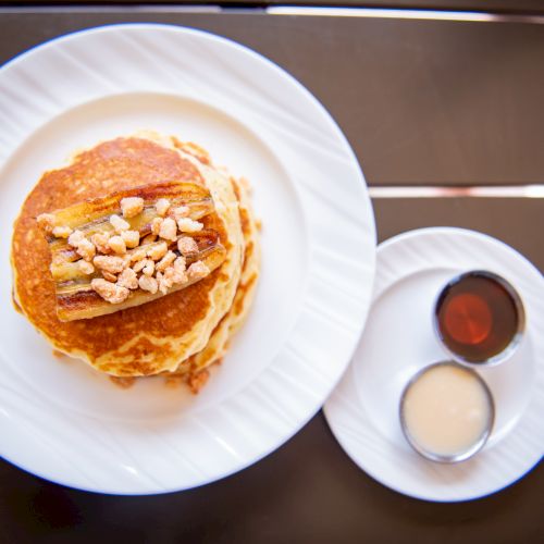A stack of pancakes topped with nuts on a plate, served with syrups on the side on a smaller plate.