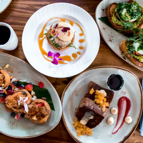 An assortment of dishes, including avocado toast, pancakes with fruit, a dessert, and coffee, is arranged on a wooden table.