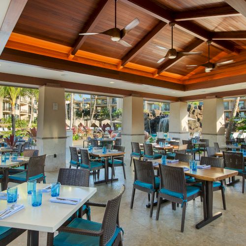 The image shows a bright restaurant with wooden ceiling, tables set with blue glasses, and outdoor views through large windows.