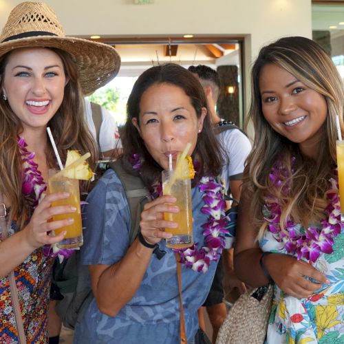 Three people are smiling, holding tropical drinks, wearing leis and summer attire, possibly at a festive or tropical-themed event.