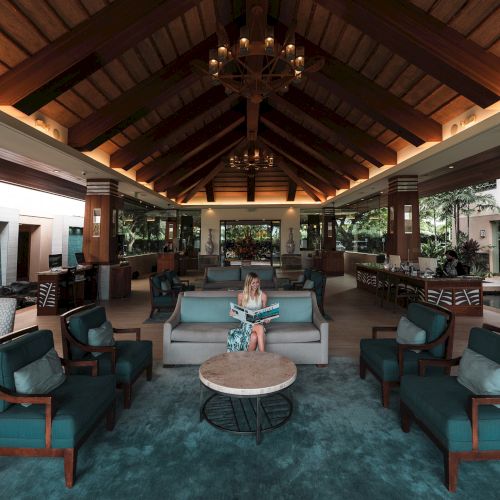 A spacious, elegant lobby with high ceilings and chandeliers, featuring comfortable seating and a person reading a magazine.
