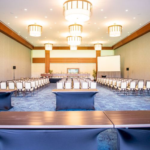 A large, well-lit conference room features rows of chairs and tables with a projector screen at the front.