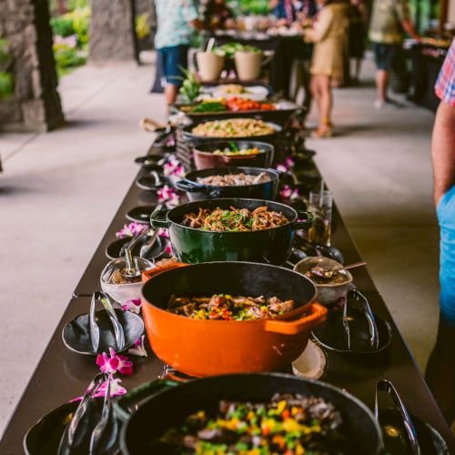 A buffet-style setup with various colorful dishes in pots on a long table, adorned with purple flowers, in an outdoor venue.