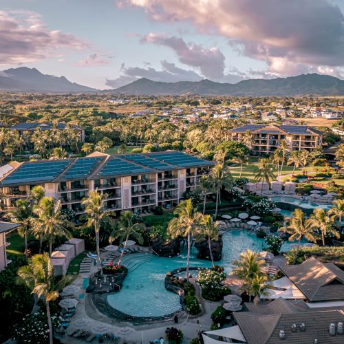 The image shows a resort with pools, palm trees, and surrounding buildings, set against a backdrop of mountains and a partly cloudy sky.