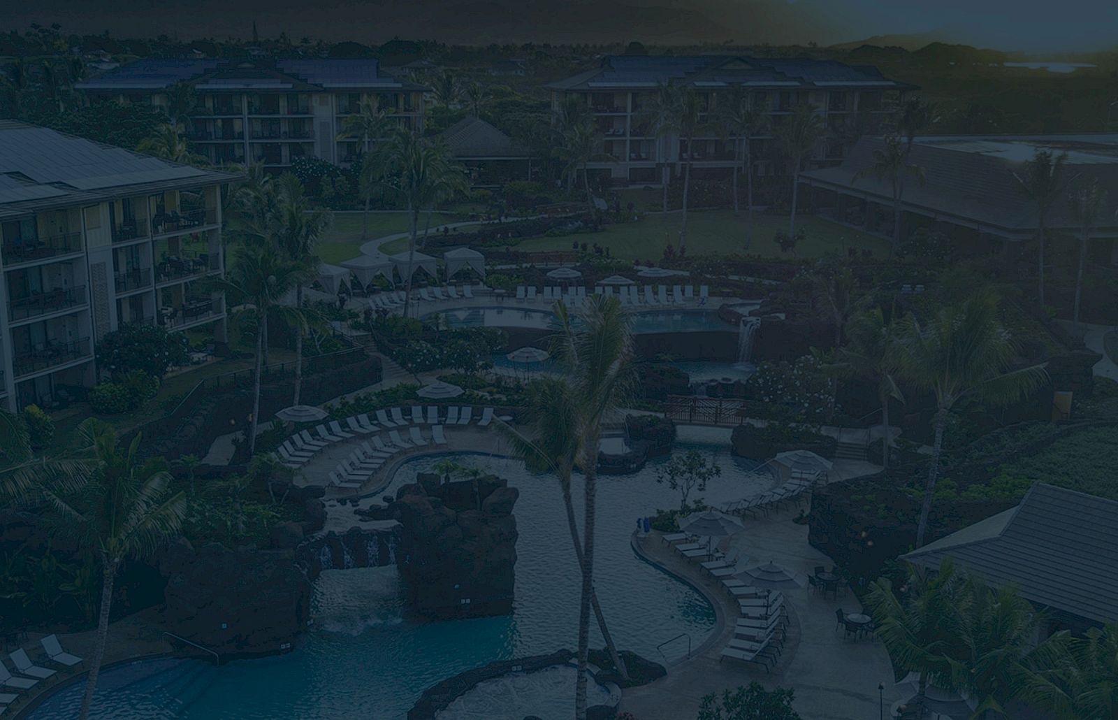The image shows a resort with multiple pools, lounge chairs, palm trees, and surrounding buildings, captured from an elevated viewpoint.