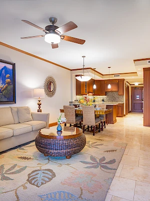 A well-lit living room with a sofa, TV, coffee table, ceiling fan, and kitchen area. Decor is warm and inviting, featuring a large wall painting.