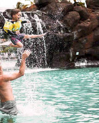 A man in a pool throws a child into the air, with a waterfall and rocks in the background.