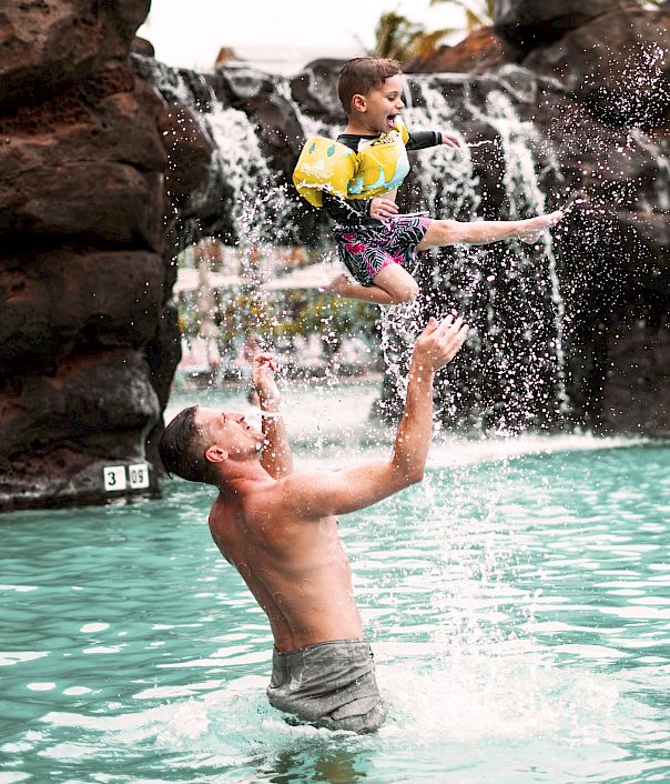 A man in a pool throws a child into the air, with a waterfall and rocks in the background.
