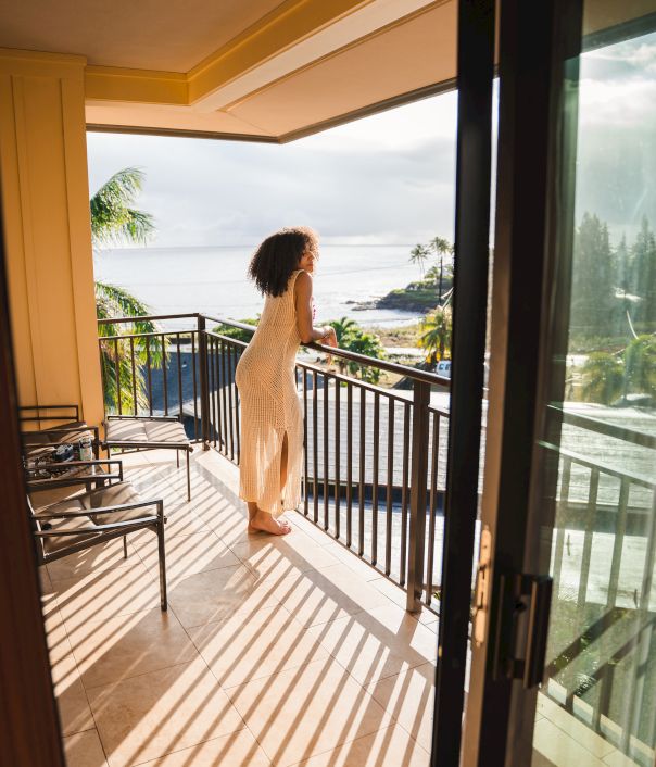 A person in a dress stands on a balcony, gazing at the ocean view, with tropical surroundings and sunlight casting shadows.