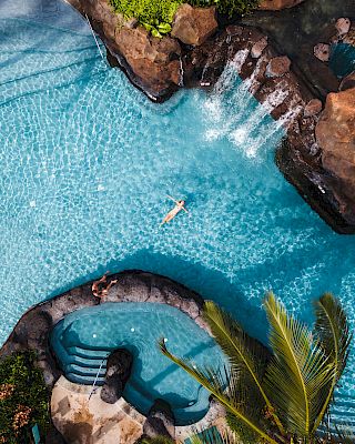 Aerial view of a pool with a person swimming, surrounded by rocks and waterfalls, adjacent to a hot tub area with a palm tree nearby.