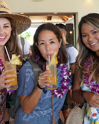 Three people wearing leis are smiling and holding tropical drinks with pineapple garnishes indoors.