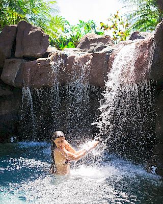 A person is enjoying a waterfall in a tropical setting, surrounded by rocks and palm trees, wearing swimwear and smiling in the water.