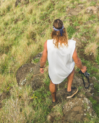 A person with long hair stands on a grassy cliff edge, wearing a casual outfit and holding a hat, overlooking a lush, green landscape below.