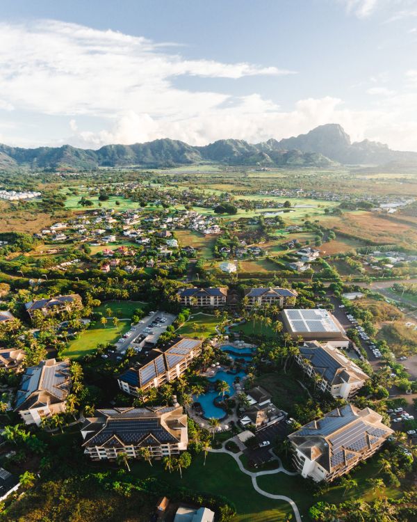 Aerial view of a resort or village with buildings, a central pool, and lush green landscape surrounded by hills and fields, under a clear sky.