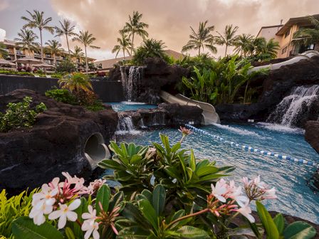 A resort pool with cascading waterfalls, tropical plants, and pink flowers in the foreground at sunset. Tone: tranquil resort vibes.